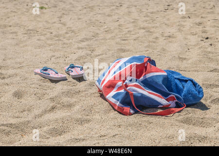 Union Jack Strandtasche & Flip-Flops am Sandstrand. Für 2021 Aufenthalte in Großbritannien, Urlaub zu Hause, Aufenthalt in Cornwall, Badeurlaub, Flip-Flop-Schuhe. Stockfoto