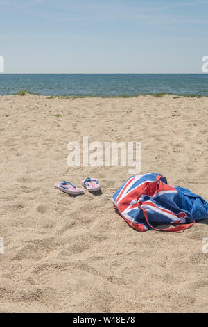 Union Jack Strandtasche & Flip-Flops am Sandstrand - für 2021 Aufenthalt in Großbritannien, Urlaub zu Hause, Aufenthalt in Cornwall, Badeurlaub, Flip-Flop-Schuhe. Stockfoto