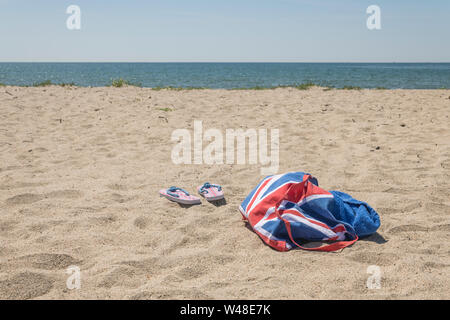 Union Jack Strandtasche & Flip-Flops am Sandstrand. Für 2021 Aufenthalte in Großbritannien, Urlaub zu Hause, Aufenthalt in Cornwall, Badeurlaub, Flip-Flop-Schuhe. Stockfoto