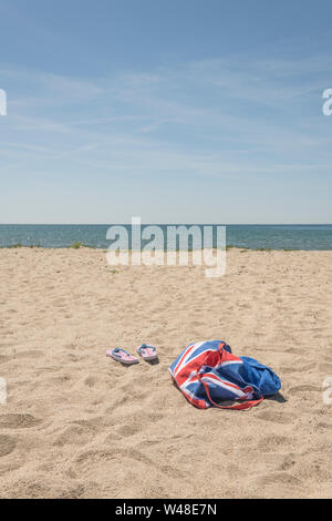 Union Jack Strandtasche & Flip-Flops am Sandstrand. Für 2021 Aufenthalte in Großbritannien, Urlaub zu Hause, Aufenthalt in Cornwall, Badeurlaub, Flip-Flop-Schuhe. Stockfoto