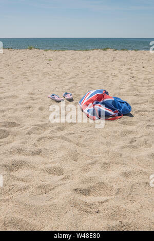 Union Jack Strandtasche & Flip-Flops am Sandstrand. Für 2021 Aufenthalte in Großbritannien, Urlaub zu Hause, Aufenthalt in Cornwall, Badeurlaub, Flip-Flop-Schuhe. Stockfoto