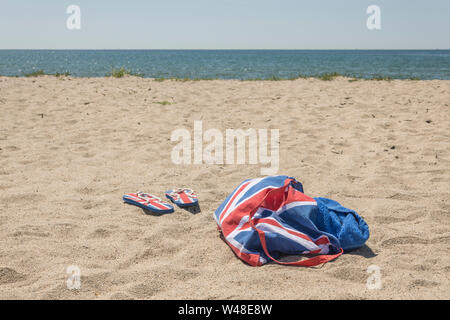 Union Jack Strandtasche & Flip-Flops am Sandstrand - für 2021 Aufenthalte in Großbritannien, Urlaub zu Hause, Aufenthalt in Cornwall, Badeurlaub, Flip-Flop-Schuhe. Stockfoto