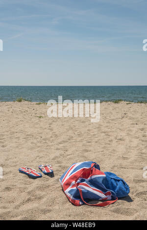 Union Jack Strandtasche & Flip-Flops am Sandstrand. Für 2021 Aufenthalte in Großbritannien, Urlaub zu Hause, Aufenthalt in Cornwall, Badeurlaub, Flip-Flop-Schuhe. Stockfoto