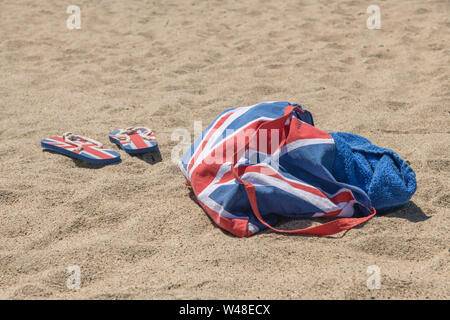 Union Jack Strandtasche & Flip-Flops am Sandstrand. Für 2021 Aufenthalte in Großbritannien, Urlaub zu Hause, Aufenthalt in Cornwall, Badeurlaub, Flip-Flop-Schuhe. Stockfoto