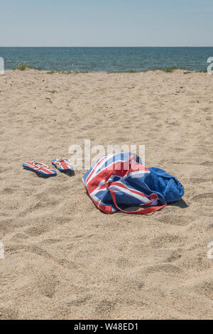 Union Jack Strandtasche & Flip-Flops am Sandstrand. Für 2021 Aufenthalte in Großbritannien, Urlaub zu Hause, Aufenthalt in Cornwall, Badeurlaub, Flip-Flop-Schuhe. Stockfoto