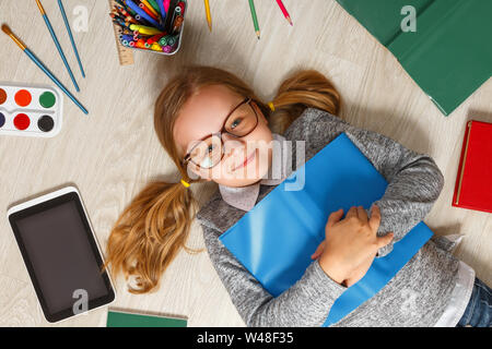 Süße kleine Mädchen in Gläsern mit einem Buch auf dem Boden liegt. Ein Kind ist von einem Buch, Tablet, Farben, Pinsel, Stifte umgeben. Stockfoto