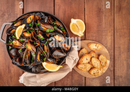 Marinara Muscheln, Moules Mariniere, mit geröstetem Brot und Zitronenscheiben, in einem Kochtopf, Schuß von der Oberseite in einem dunklen Holzmöbeln im Landhausstil Hintergrund Stockfoto