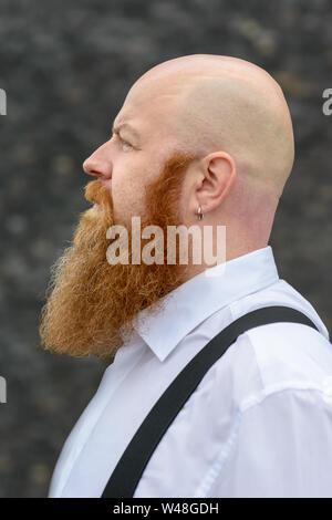 Profil anzeigen von einem bärtigen rothaarigen Mann mit rasiertem Kopf tragen Ohrring und geschweifte Klammern über einen grauen Hintergrund Stockfoto
