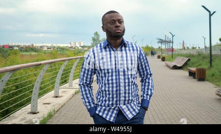 Schwarze Kerl in Plaid Shirt und Jeans auf der Straße. Junge afro-amerikanischer Mann ist zu Fuß in der City Park in trüben Sommertag. Er faltete die Hände in den Taschen. Er schaut besorgt und ängstlich. Stockfoto