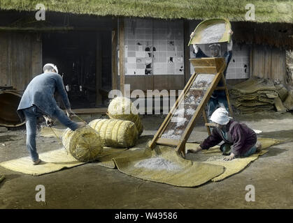 [1920s Japan - Japanische Bauern Worfeln Reis] - Eine weibliche Landwirt entfernt Kleie von Reis in ein trennender Maschine durch einen männlichen Landwirt. Ein anderer Mann Landwirt Verpackung Reis ballen. 20. Jahrhundert vintage Glas schieben. Stockfoto