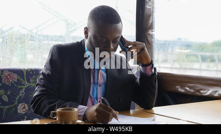 Schwarze Kerl spricht auf dem Smartphone. Afro-amerikanische Geschäftsmann ruft auf dem Handy im Sommer Hütte Cafe sitzen. Er trinkt eine Tasse Kaffee am Tisch. Er trägt Hemd und Anzug Jacke. Stockfoto