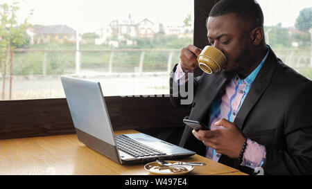 Afro-amerikanische Mann hat Frühstück. Schwarzer Geschäftsmann arbeitet auf Laptop mit Smartphones im Café und trinkt Kaffee. Er tippt auf computer handy Bildschirm schaut. Trägt in Hemd und Anzug Jacke. Stockfoto