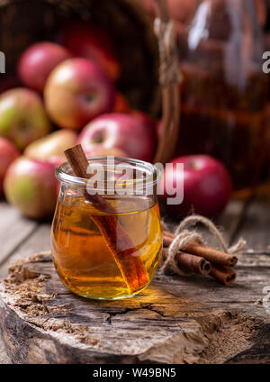Glas Apfelwein mit Zimtstangen auf einem rustikalen Oberfläche mit roten Äpfeln im Hintergrund Stockfoto