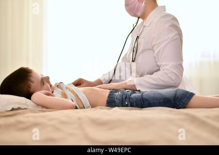 Ein Arzt hört mit einem Stethoskop den Herzschlag und Atem eines lächelnden Jungen. Das Baby ist zu Hause auf dem Bett, der Arzt sitzt in der Nähe. Stockfoto