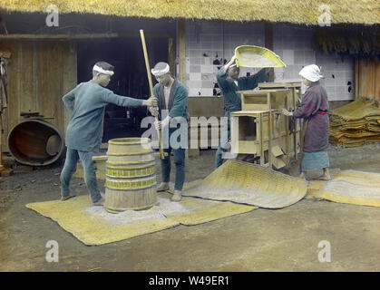 [1920s Japan - Japanische Bauern Worfeln Reis] - Japanische Landwirte sind Schälen (vorne) und Worfeln Reis (hinten). Die Mühle in der Front ist aufgerufen, eine dozurusu (土摺臼) und die WORFELN Maschine im Rücken ist ein toumi (唐箕). 20. Jahrhundert vintage Glas schieben. Stockfoto