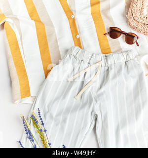 Komfortable gestreifte Bluse, blaue Hosen, Stroh Hut, Sonnenbrille und wilden Feldblumen auf weißem Hintergrund. nach oben Foto anzeigen, pastell Farben in der Kleidung, g Stockfoto