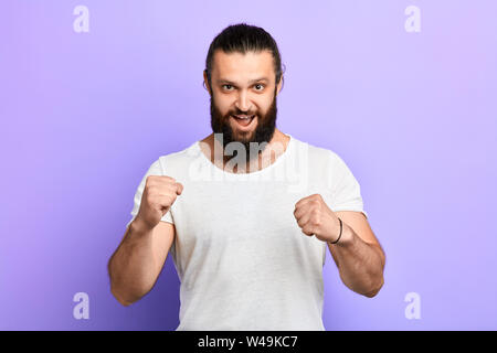Positive junger Mann mit weit geöffneten Mund tragen stilvolle Kleidung mit geballten Fäusten in Studio posieren. aufgeregt Mann, der bereit ist zu kämpfen, Sport, Freizeit, Stockfoto