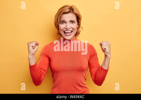 Gerne nette charmante junge Frau mit toothy Lächeln erhobenen Hände und feiert Erfolg. Bis pjrtarit schließen. Wir haben es geschafft. glück Konzept Stockfoto