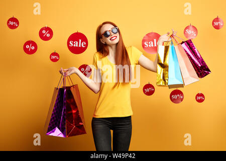 Fröhlicher, lustiger rothaarige Frau in Sonnenbrille mit bunten Einkaufstaschen auf gelbem Hintergrund. nach oben Porträt schließen. Lifestyle, Hobby Stockfoto