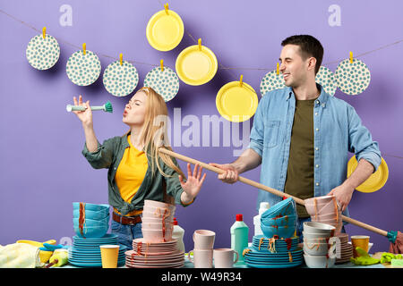 Schöne blonde Frau, die vorgibt, Song mit Pinsel und lächelnden jungen Mann spielt mit Mop wie Gitarre in der Küche mit blauen Wand zu singen. Stockfoto