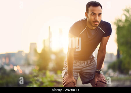 Portrait von aktiven tausendjährigen Mann Joggen in der Dämmerung mit einem urbanen Stadtlandschaft und den Sonnenuntergang im Hintergrund Stockfoto