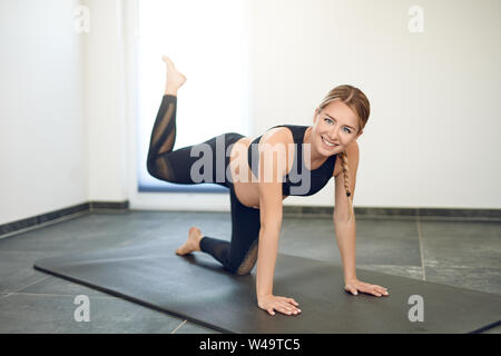 Fit gesunde junge schwangere Frau die Ausübung einer yoga Workout und Bein auf einer Matte zu Hause schauen in die Kamera mit einem glücklichen Lächeln Stockfoto