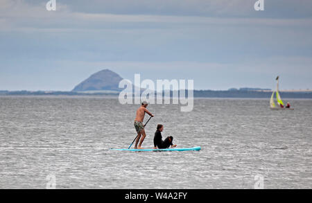 Portobello, Edinburgh, Schottland, Großbritannien. Juli 2019 21. Sportliche Aktivitäten gerade weg von Portobello Beach ein Mann Stand up Paddle Boarding mit weiblichen Passagier mit Berwick Gesetz und in einem Beiboot hinter sich. Stockfoto