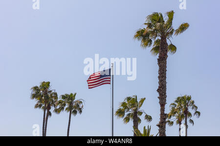 USA Symbol. Vereinigte Staaten von Amerika Flagge auf einer Stange winken, Palmen und dem klaren, blauen Himmel Hintergrund. Stockfoto