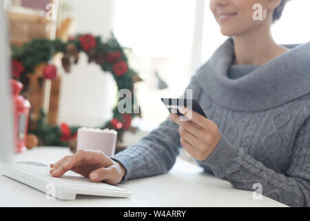 Close-up zufrieden junge Frau im grauen pullover Sitzen am Tisch und Eingabe von Kreditkarteninformationen beim Zahlen für on-line-Kauf Stockfoto