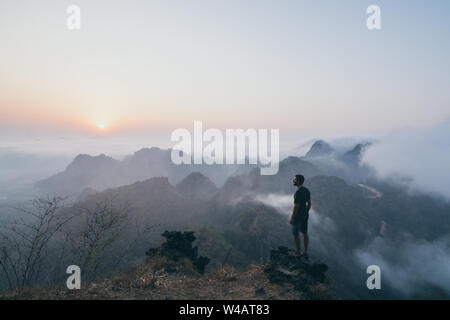 Jungen kaukasischen Mann stand auf dem Felsen mit Blick auf den tropischen Bergen bei Sonnenaufgang nebligen Morgen in Hpa-an, Myanmar Stockfoto