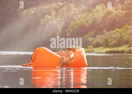 Eine orange, Sofa oder eine aufblasbare Liege schwimmt den Fluss entlang, in dem ein junger bärtiger Mann der kaukasischen Ethnie schläft, eco - Tourismus in einem moun Stockfoto