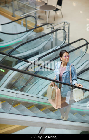 Lächelnd modische junge Dame in rosa Sonnenbrille auf der Rolltreppe in Mall und Holding Shopping Taschen Stockfoto