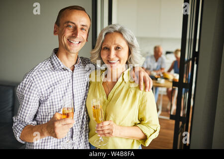 Taille bis Portrait von Happy reifes Paar holding Champagner Flöte an der Kamera zu Hause mit Freunden im Hintergrund posiert Suchen, Kopieren Raum Stockfoto
