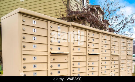 Panorama rahmen Wohn- Mailboxen mit nummerierten Fächer an der Seite einer Straße. Wohnungen, schneebedeckte Berge und bewölkter Himmel können in gesehen werden. Stockfoto