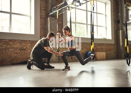 Tun hocke Übung. Selbstbewussten jungen Personal Trainer ist, schlanke sportliche Frau, wie Kniebeugen mit Trx fitness Bänder zu tun beim Training im Fitnessstudio. TRX Training. Zusammen trainieren. Aktiven Lebensstil Stockfoto