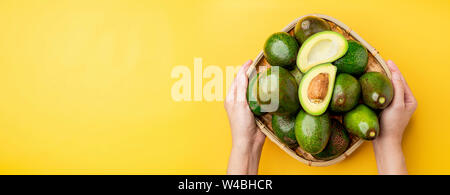 Hand, die biologischen Avocados halbieren mit Saatgut und ganze Früchte im Korb auf der gelben Tabelle Hintergrund. gesund Super Foods für Ernährung. Frisches Gemüse her Stockfoto