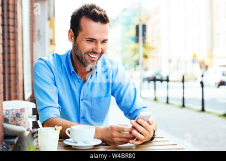Glückliche junge Mann im Internet surfen auf dem Smartphone im Café draußen sitzen. Stockfoto