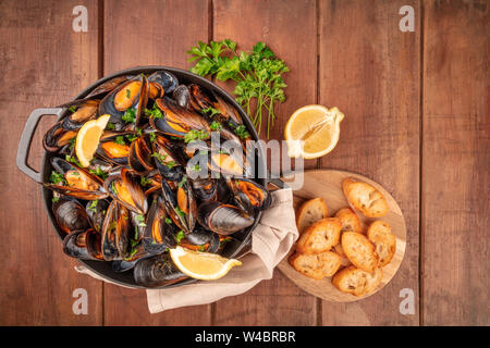 Marinara Muscheln, Moules Mariniere, mit geröstetem Brot, Petersilie, Zitronenscheiben, im Kochtopf, Overhead shot auf einem dunklen Holzmöbeln im Landhausstil Hintergrund Stockfoto