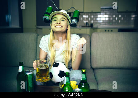 Junge Frau gucken Fußball Spiel im tv in der Nacht. Fröhliches positives Modell lächelnd. Hand in Hand. Sie suchen. Glas Bier und Flaschen auf dem Tisch. Stockfoto