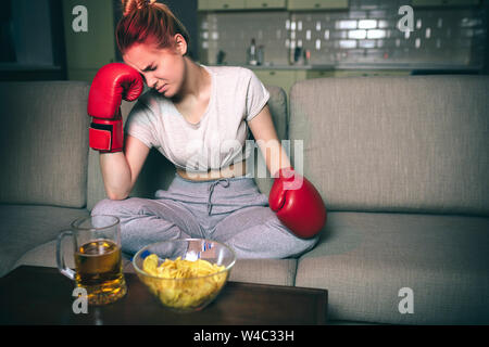 Junge Frau watch Boxen im tv in der Nacht. Verärgert unzufrieden Modell sitzen auf dem Sofa und halten Sie den Kopf auf die Hand. Das Tragen der roten Sport Boxhandschuhe. Allein im dunklen Zimmer. W Stockfoto