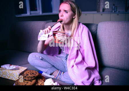 Junge Frau ansehen Film in der Nacht. Das Sitzen auf einem Sofa im Zimmer allein. Eis Essen mit dem Löffel. Schreien. Traurigen Film oder TV-Serien. Dunkle Zimmer Stockfoto