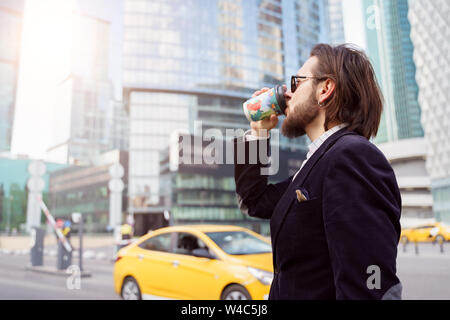 Foto an der Seite des jungen Brunet mit Bart und Ohrring im Ohr das Trinken aus Glas in der Stadt auf dem Hintergrund von Hochhäusern, gelbe Auto Stockfoto