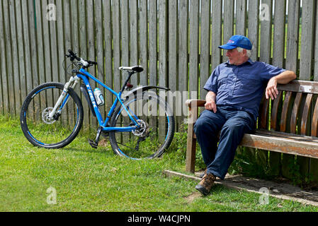 Älterer Mann sitzt auf der Bank, Fahrradverleih, lehnte sich gegen Zaun, England Großbritannien Stockfoto