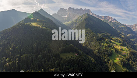 Luftbilder über eine Alpine Valley und üppig grüne Pinienwälder mit Chalets und hohen Berge, die das Tal. Stockfoto