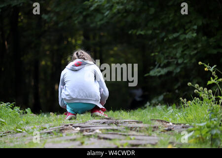 Kleine Mädchen spielen auf dem Weg in einen dunklen Wald, Ansicht von hinten. Kind hocken sammelt Pflanzen im Sommer Stockfoto