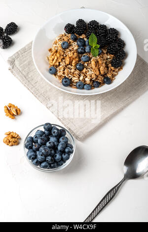 Gesundes Frühstück. Hausgemachtes Müsli, Müsli, Cerealien mit Brombeeren, Heidelbeeren, Nüsse, Honig und Minze in einer weißen Schüssel auf einem weißen Hintergrund. Stockfoto