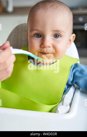 Portrait Of Smiling Baby Boy zu Hause in Hochstuhl Zugeführtes feste Nahrung von der Mutter mit Löffel Stockfoto