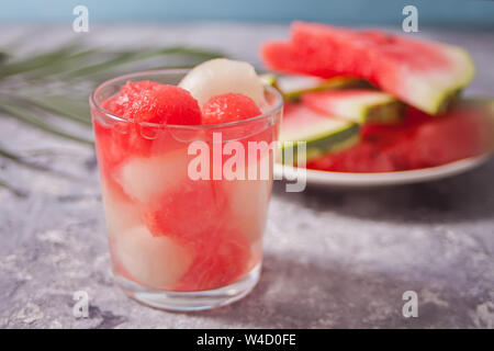 Obstsalat mit Melone und Wassermelone Kugeln aus Glas und Stück Wassermelone unter dem palm leaf auf dem concreate Hintergrund Stockfoto