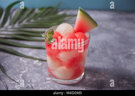 Obstsalat mit Melone und Wassermelone Kugeln aus Glas und Stück Wassermelone unter dem palm leaf auf dem concreate Hintergrund Stockfoto