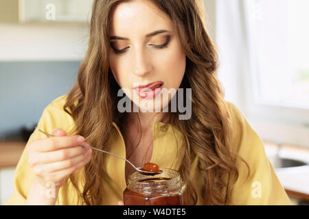 Junge schöne Mädchen ist das Frühstück zu Hause in der Küche. Sie trinkt ihren Kaffee am Morgen und mit großer Freude isst Marmelade aus der Dose. Stockfoto